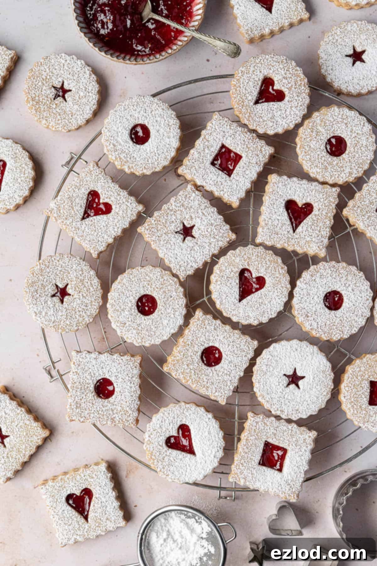 Vegan Linzer cookies arranged on a round wire cooling rack, with a bowl of raspberry jam, a sieve for icing sugar, and various cookie cutters in the background.