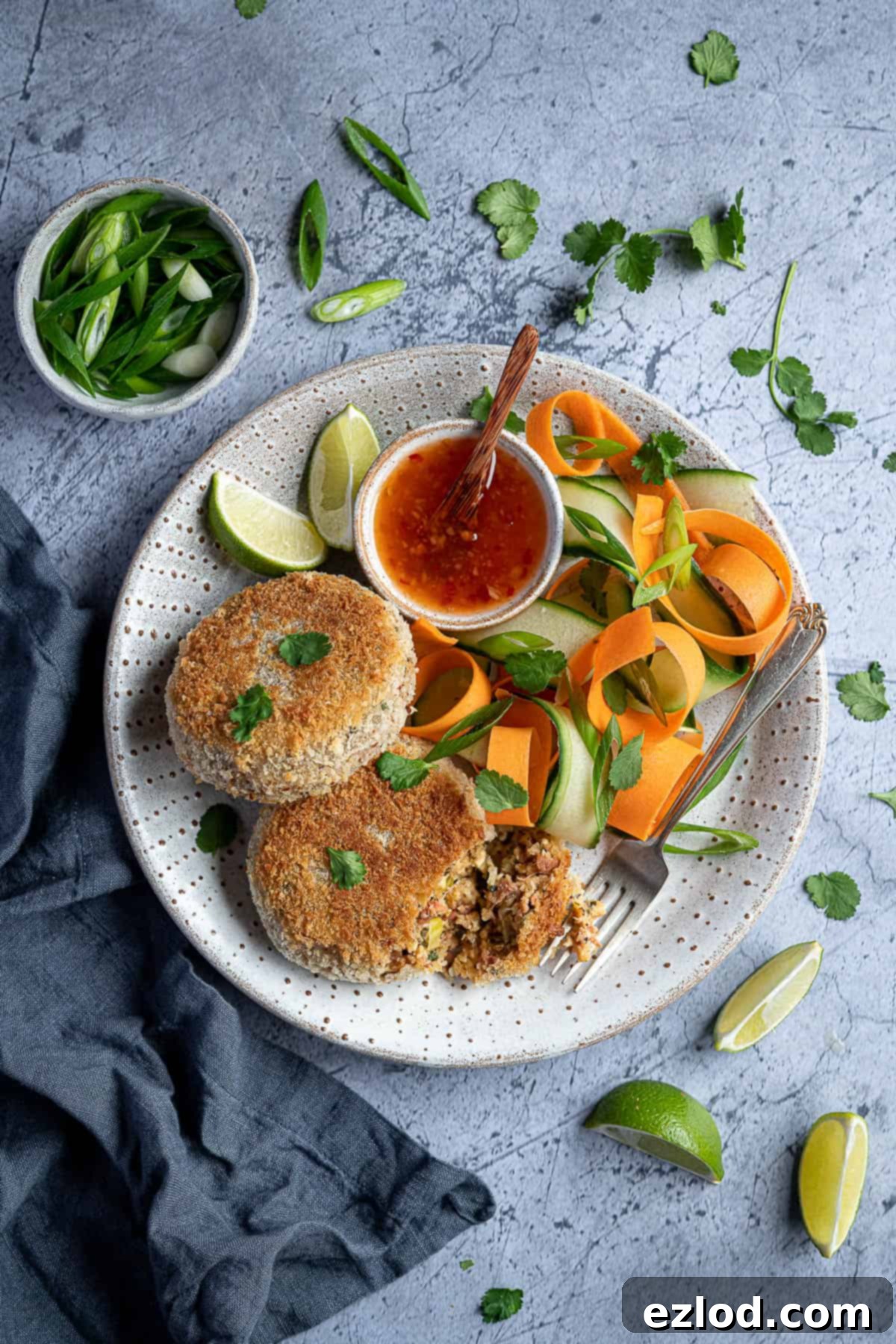 Two golden-brown vegan Thai red curry 'fish' cakes served on a white plate alongside fresh green vegetable ribbons and a small bowl of sweet chili sauce. The plate is garnished with bright lime slices, finely sliced spring onions, and vibrant fresh coriander leaves, showcasing a beautifully presented, flavorful meal.