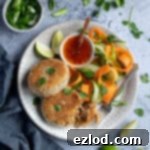 Two vegan Thai red curry 'fish' cakes on a plate with vegetable ribbons and a bowl of sweet chilli sauce, surrounded by lime slices, sliced spring onions and fresh coriander.