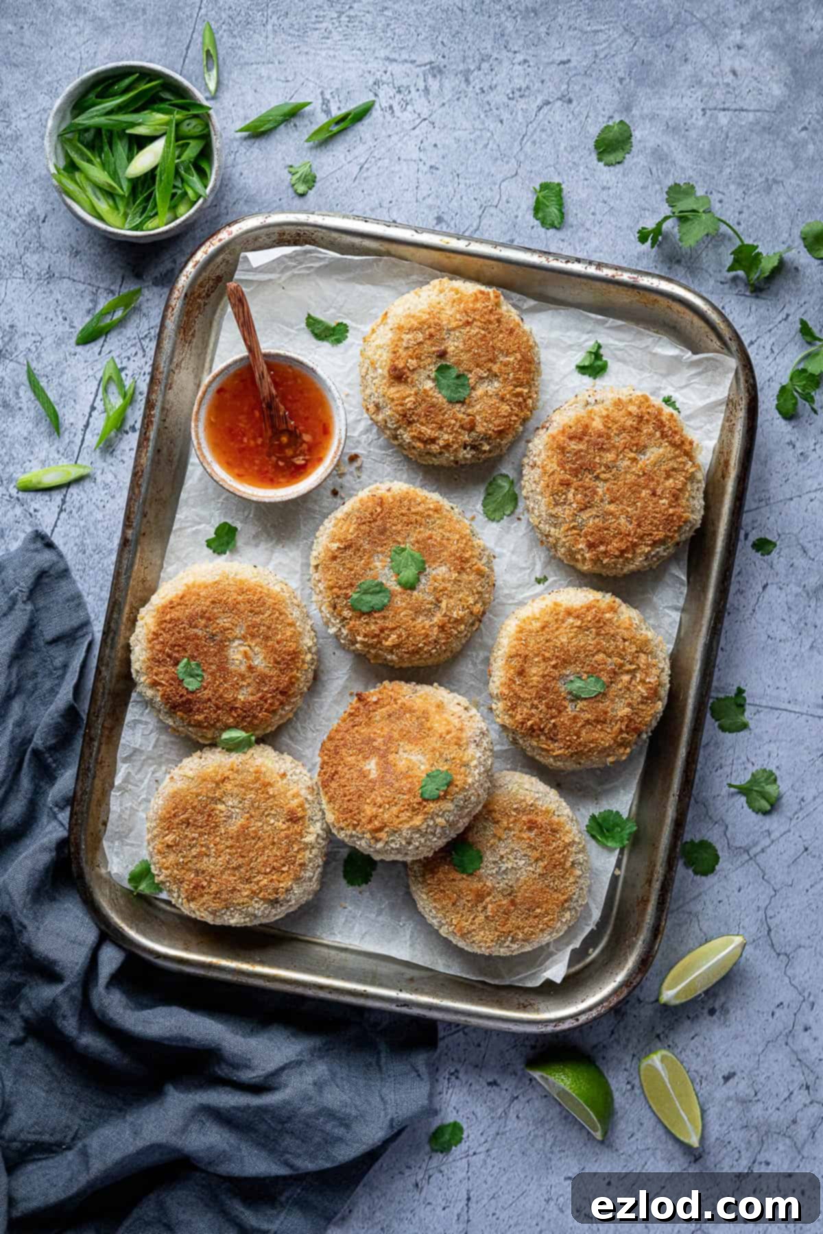 A selection of raw, coated vegan Thai red curry 'fish' cakes arranged neatly on a baking sheet, ready for cooking. Accompanying them are small bowls of sweet chili sauce and fresh garnishes like lime slices and sliced spring onions, hinting at the delicious meal to come.