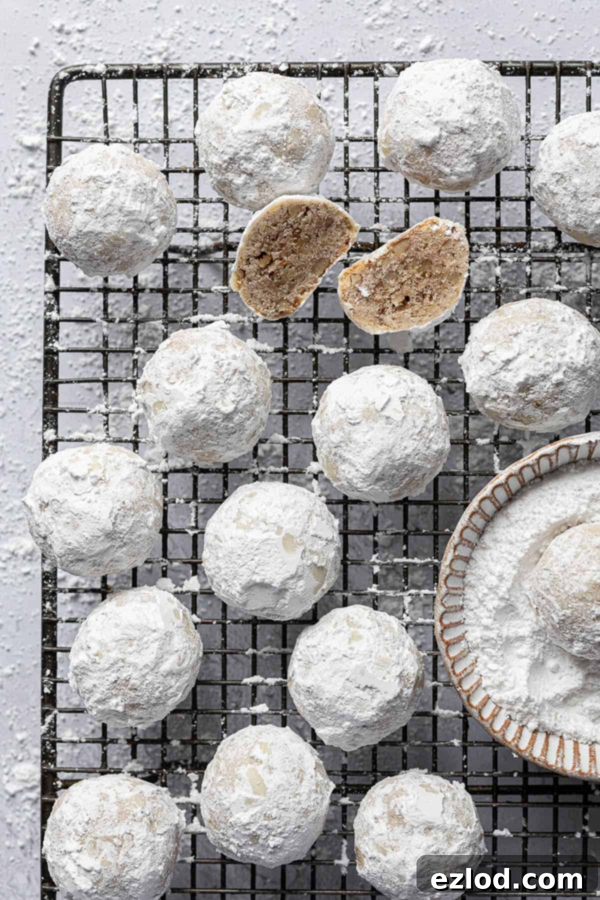 Classic Vegan Pecan Snowball Cookies 12 A collection of vegan pecan snowball cookies, some whole and one sliced in half, artfully arranged on a wire rack next to a bowl of icing sugar.
