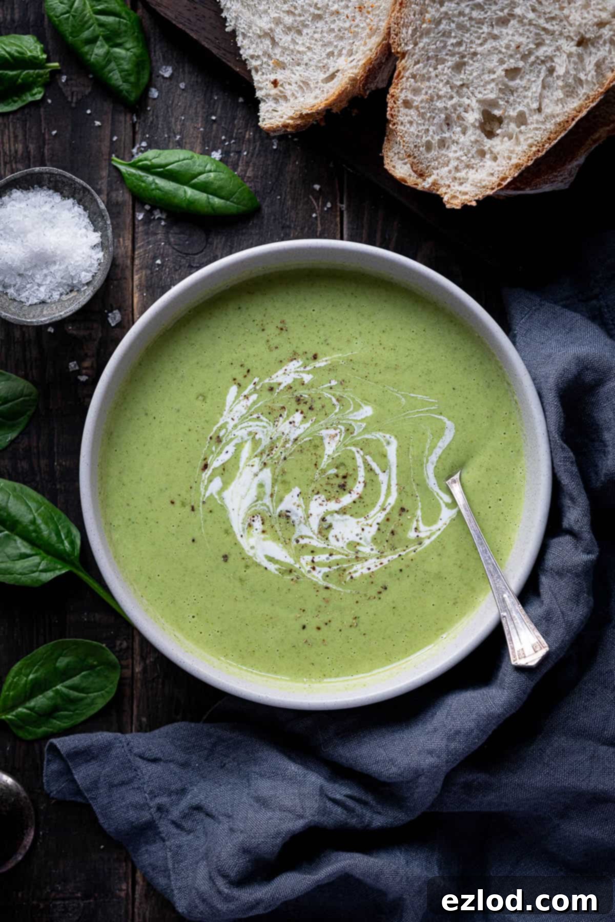 Creamy Vegan Broccoli Cheese Soup 2 A vibrant bowl of creamy cheesy vegan broccoli soup sits on a rustic wooden table, accompanied by slices of artisanal bread, a small bowl of sea salt, a soft grey linen cloth, and fresh spinach leaves, highlighting its comforting appeal.