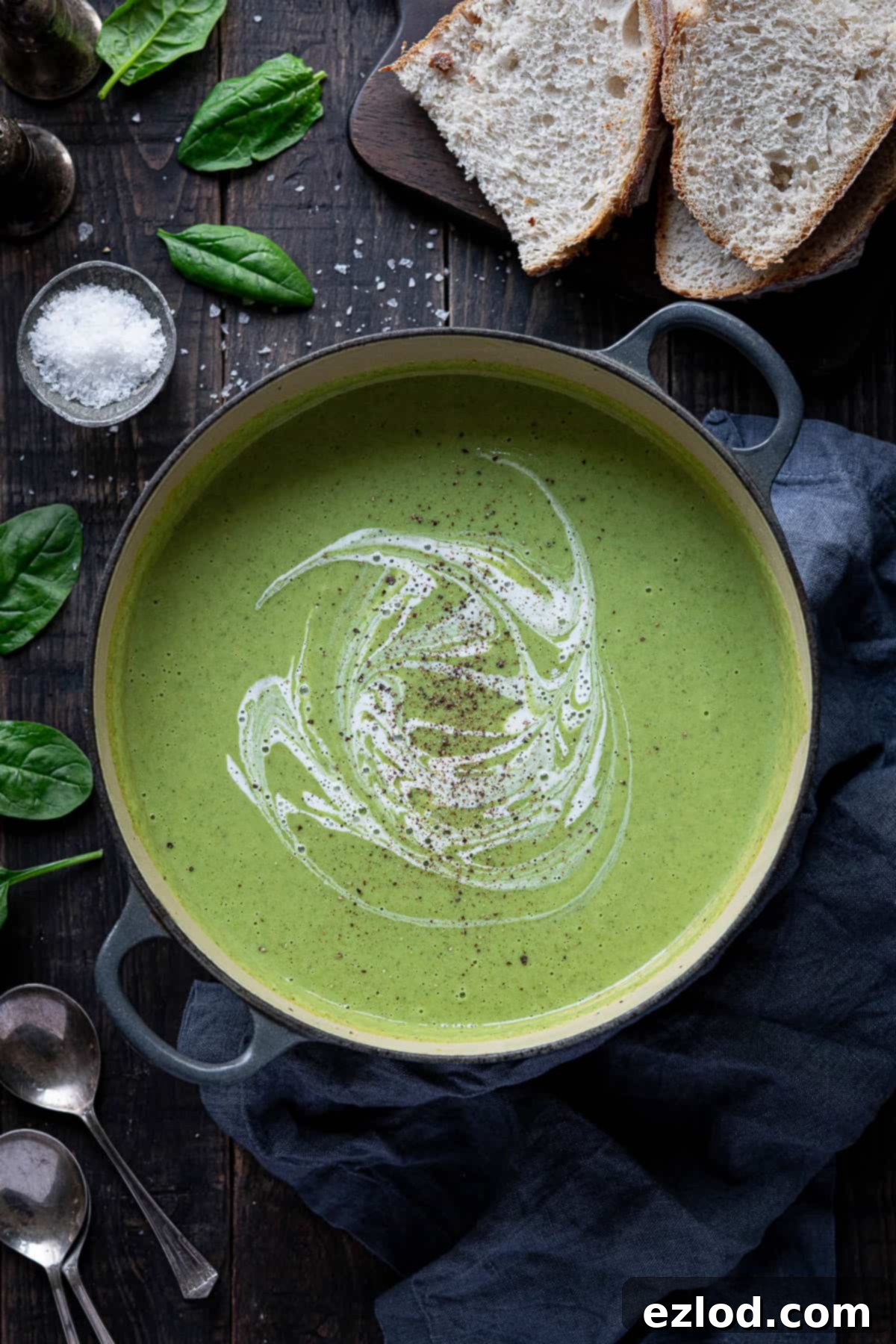 Creamy Vegan Broccoli Cheese Soup 9 A charming overhead shot of a pot of cheesy vegan broccoli soup on a wooden table, surrounded by rustic elements like bread slices, spoons, a small salt bowl, a grey cloth, and fresh spinach leaves, creating a cozy and inviting scene.