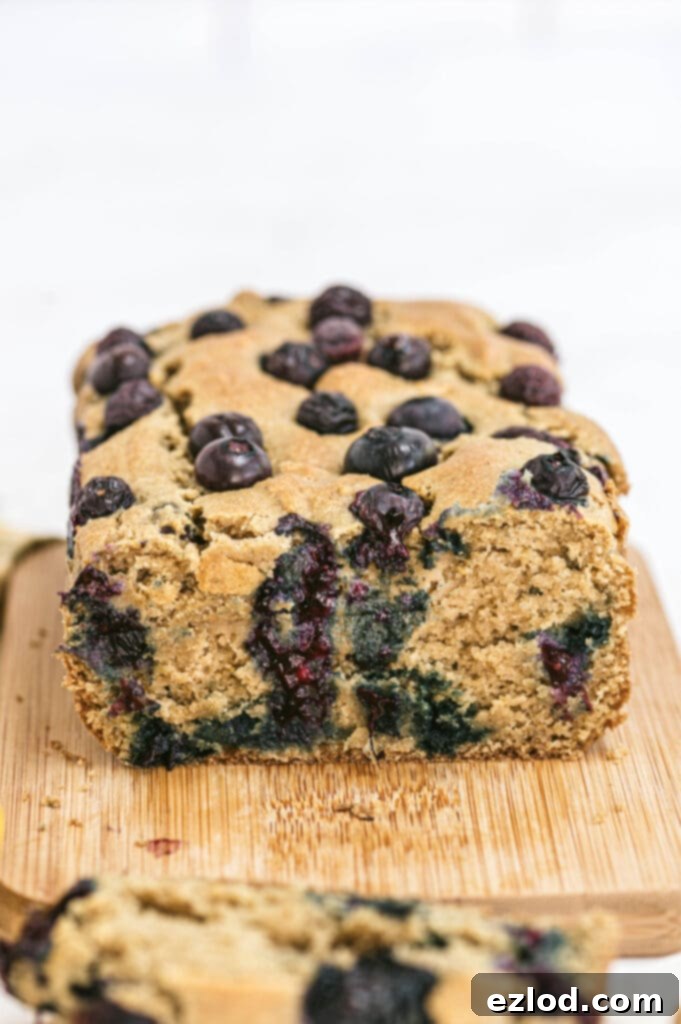 A close-up shot of a freshly baked, healthy lemon blueberry loaf resting on a rustic wooden cutting board. One slice has been carefully removed, showcasing the moist, tender crumb and the vibrant blueberries distributed throughout the bread. This inviting image highlights the loaf's perfect texture and its appeal as a wholesome treat for breakfast or a healthy snack.