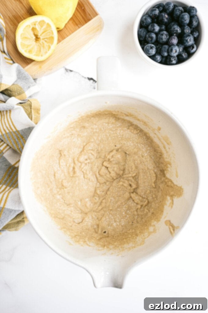An inviting overhead view of the healthy lemon blueberry bread batter in a large mixing bowl, surrounded by fresh, vibrant lemons and a bowl brimming with plump blueberries. This image perfectly captures the anticipation of baking, with all the key ingredients ready to be combined into a delicious, wholesome loaf.