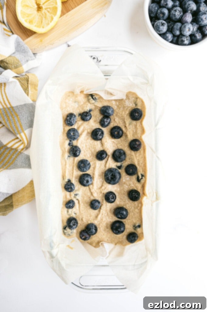 An overhead view of the healthy lemon blueberry bread batter perfectly poured into a parchment-lined loaf pan, ready to be placed in the oven. The visible blueberries are evenly distributed, hinting at the delicious treat to come.