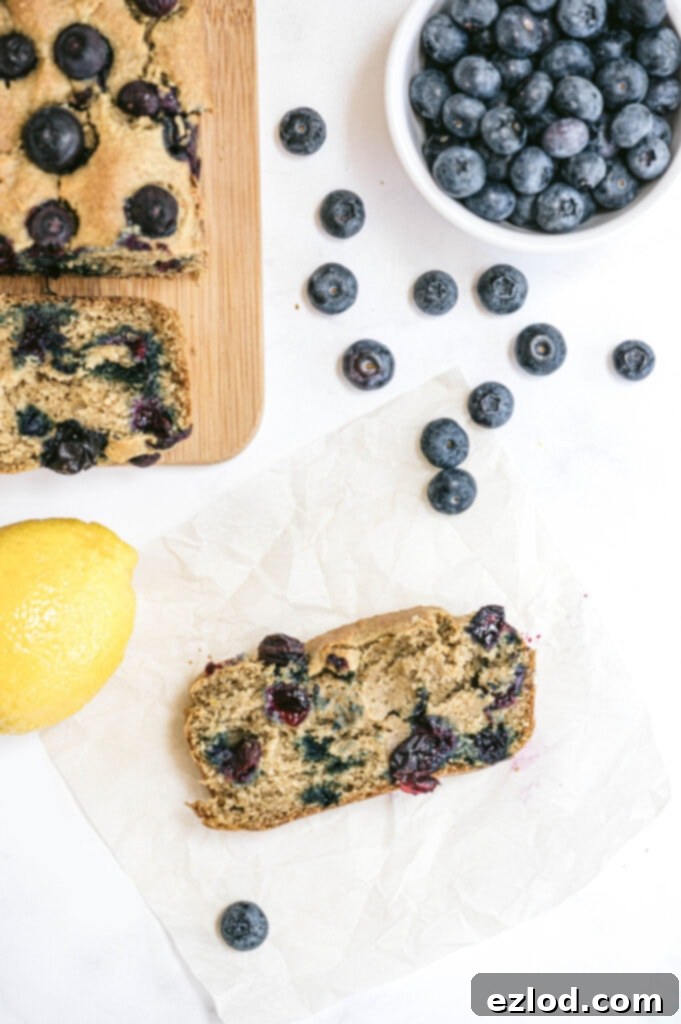 A tempting display of healthy lemon blueberry bread. A whole loaf sits on a cutting board, with a single, perfectly sliced piece resting on parchment paper. Fresh lemons and a bowl of bright blueberries surround the bread, emphasizing its natural and wholesome ingredients, perfect for a guilt-free treat.