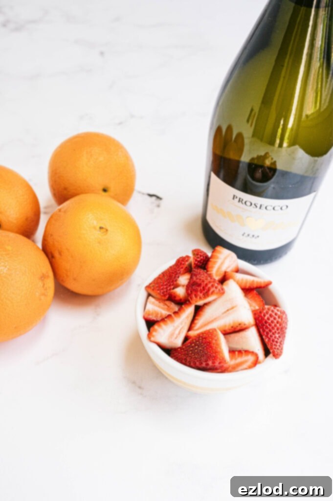 A small bowl of fresh strawberries, bright oranges, and a bottle of prosecco on a white counter, essential ingredients for mimosas