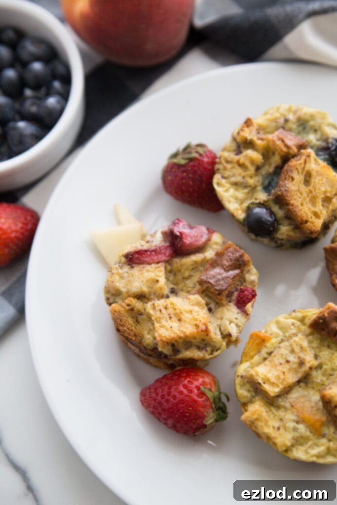 An inviting overhead photo of individual mini French Toast casserole cups, beautifully adorned with fresh fruit toppings and whole strawberries, elegantly presented on a white plate alongside additional fresh fruit.