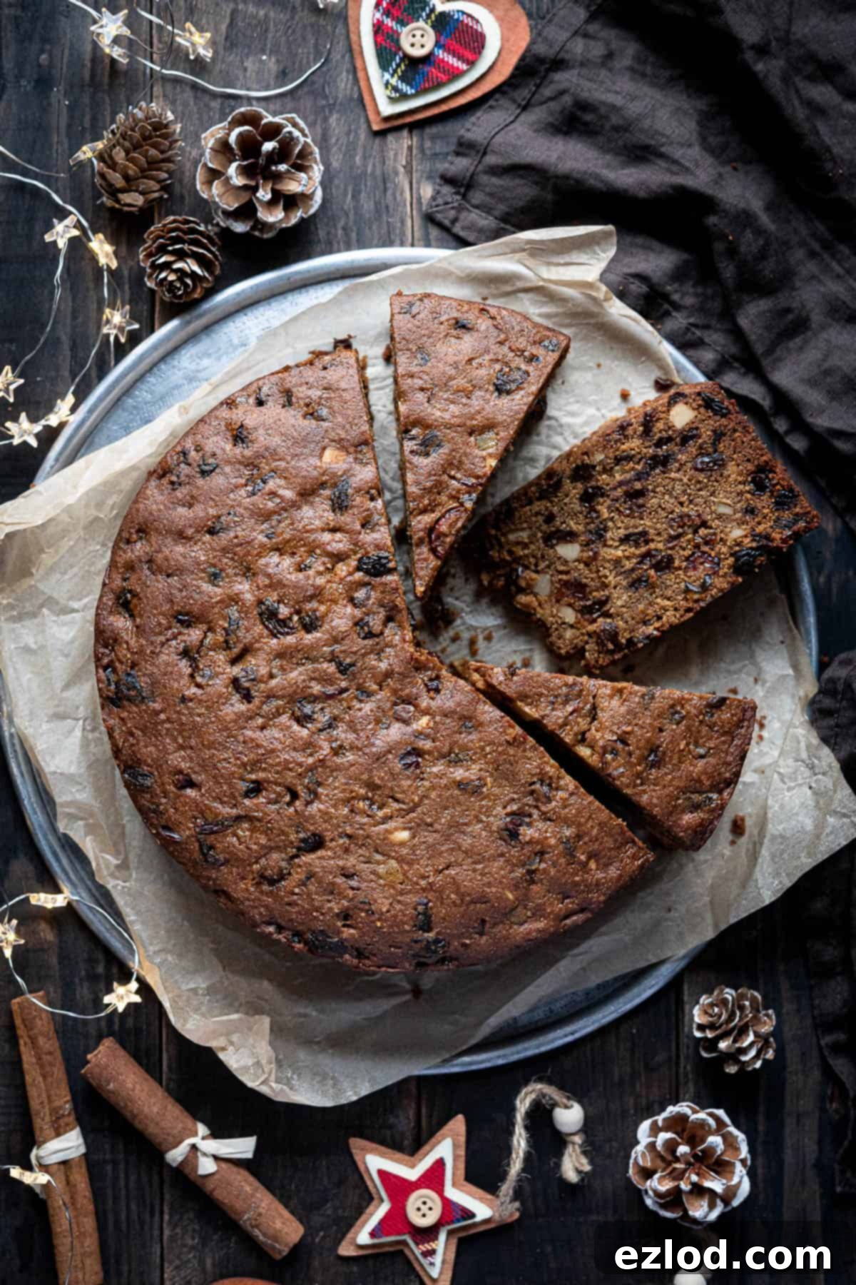 A beautifully decorated gluten-free vegan Christmas cake on a metal tray, surrounded by festive ornaments on a wooden table.