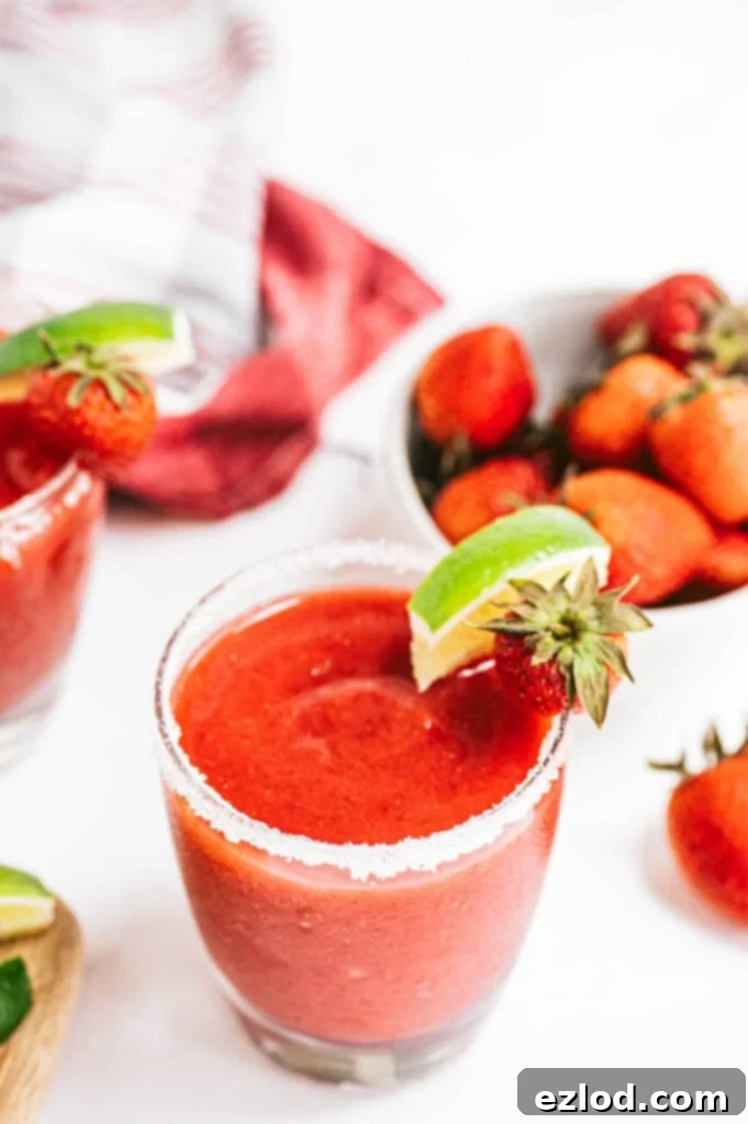 An overhead shot of a salt-rimmed glass filled with a bright pink Frozen Strawberry Rhubarb Margarita, garnished with a fresh lime wedge and a strawberry slice, resting on a rustic wooden surface under natural light.