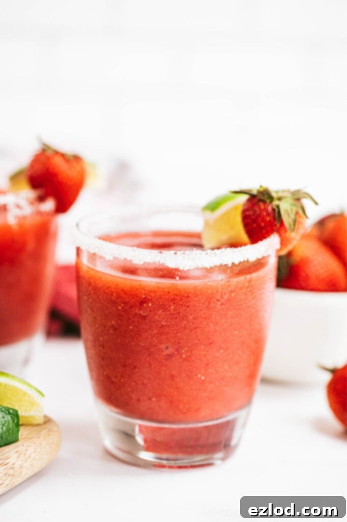 A close-up shot of a beautifully presented salt-rimmed glass, brimming with a vibrant Frozen Strawberry Rhubarb Margarita. It's garnished with a fresh lime wedge and a whole strawberry, ready for a refreshing sip.