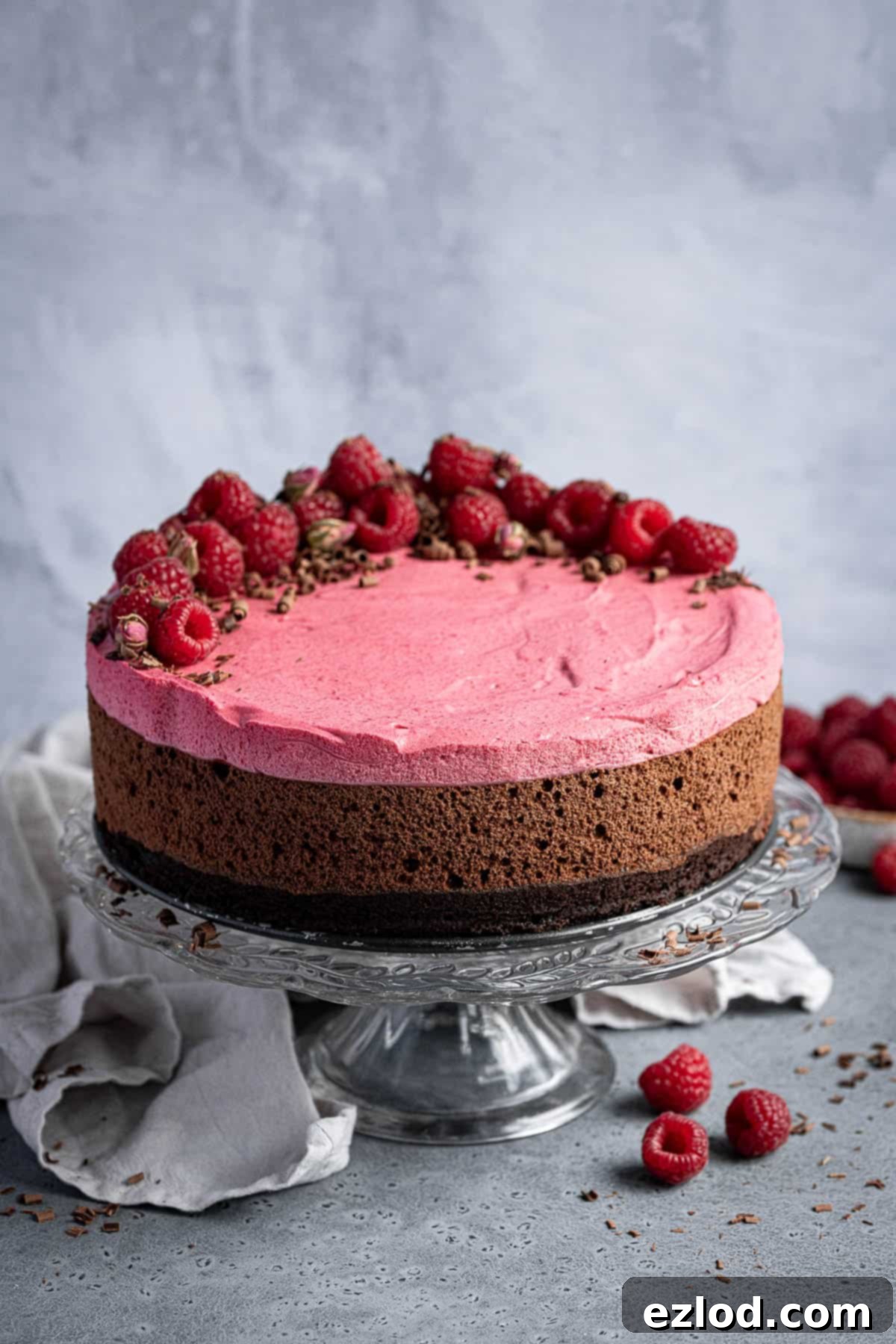 A visually striking Vegan Chocolate Raspberry Mousse Cake on a glass cake stand, garnished with fresh raspberries and a grey cloth nearby, highlighting its layers.