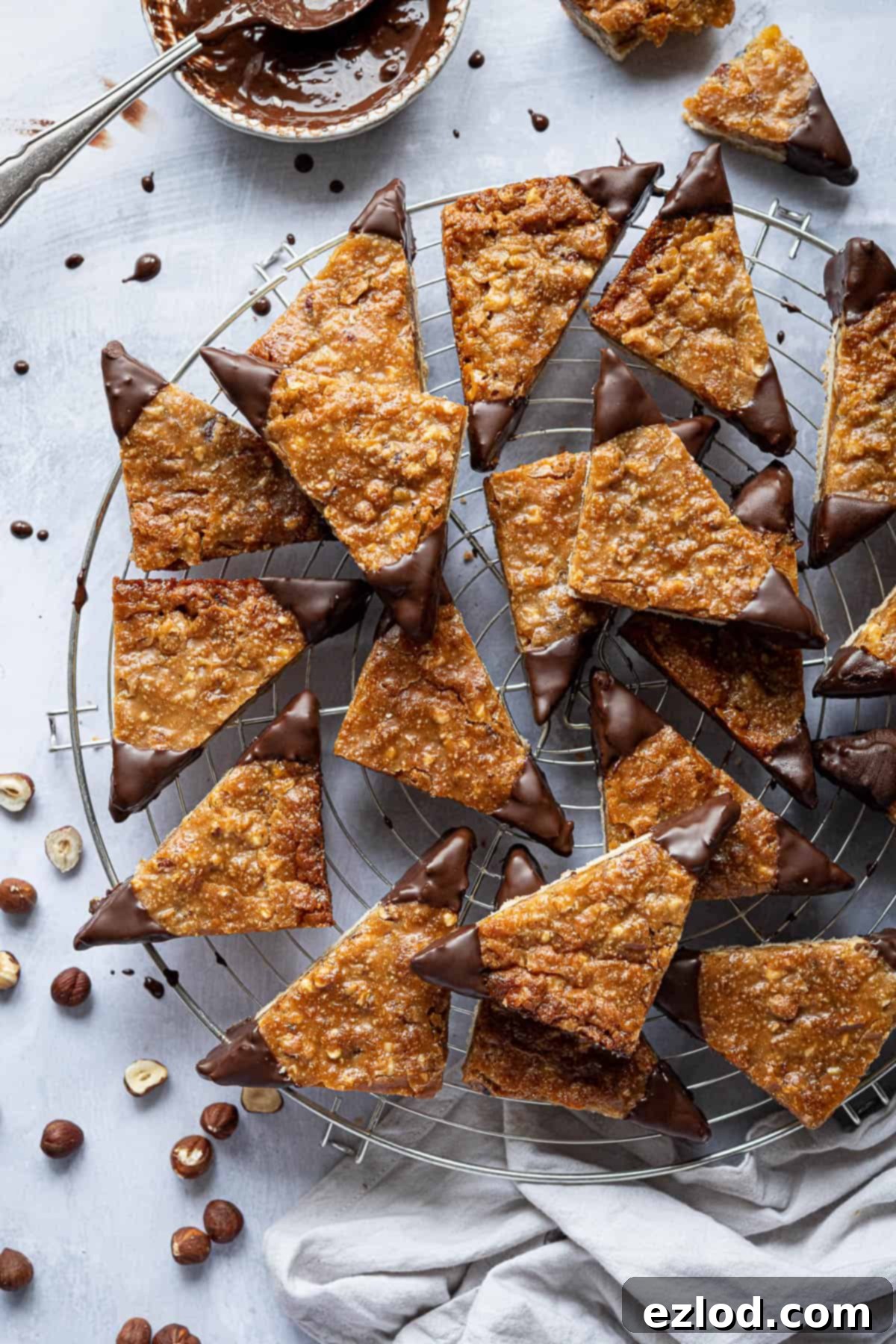Vegan nussecken on a round wire cooling rack with a bowl of chocolate and some scattered hazelnuts.