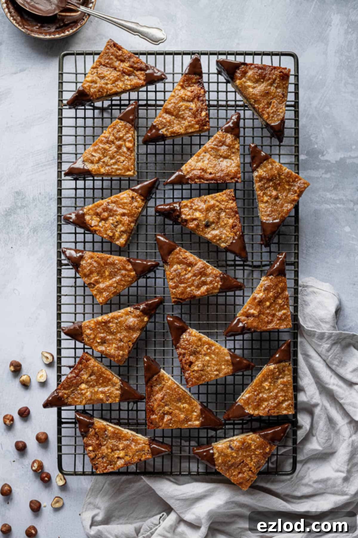 Vegan nussecken on a rectangular wire cooling rack with a bowl of chocolate, grey cloth and hazelnuts.
