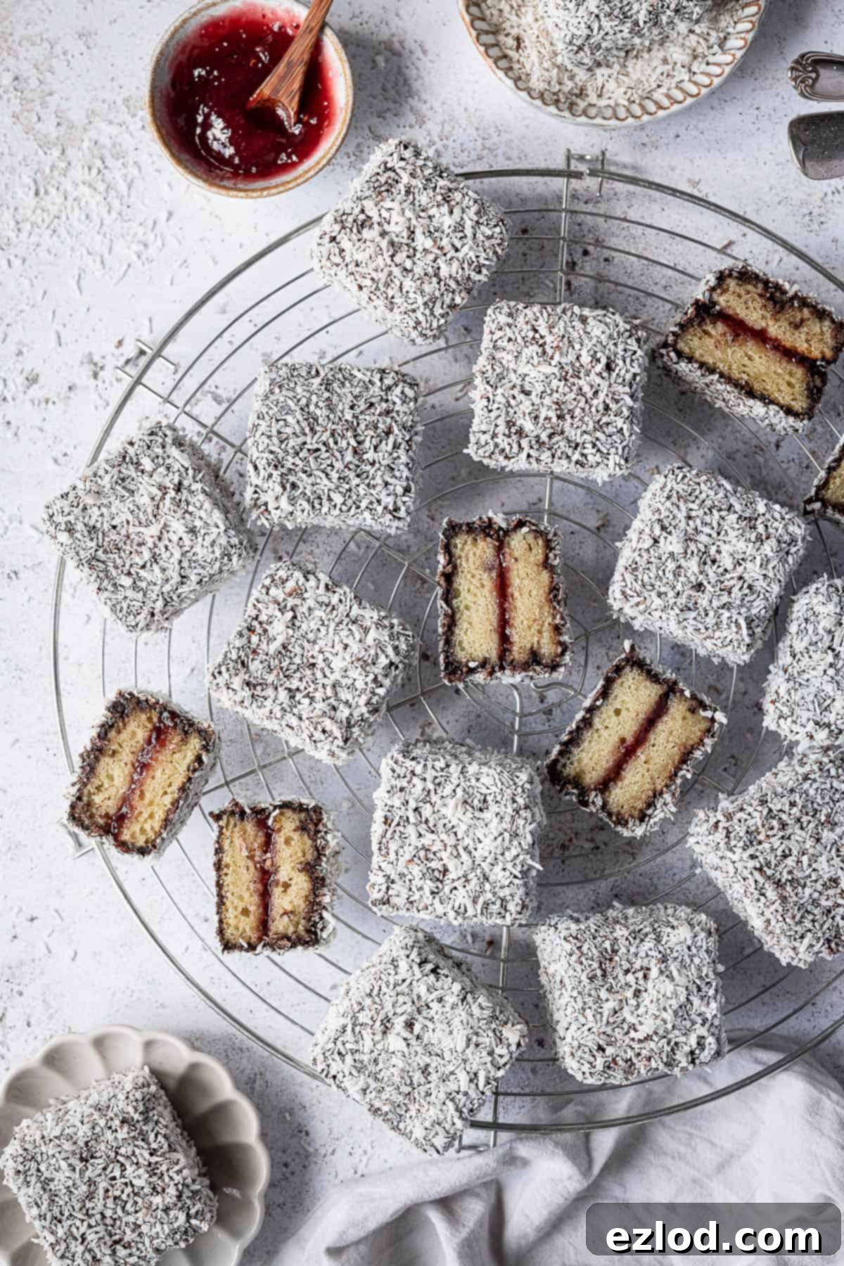Vegan Lamingtons on a round wire rack with a bowl of jam, some sliced in half.