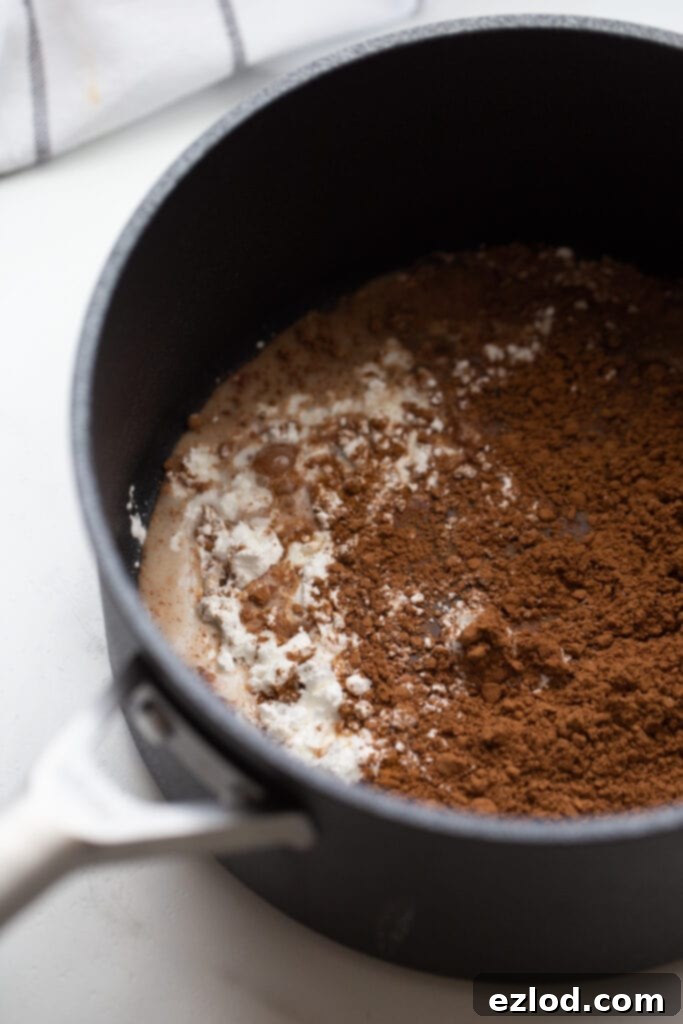 A saucepan on a stovetop containing cornstarch, cocoa powder, and a small amount of milk, ready for whisking to create the vegan chocolate pudding base.