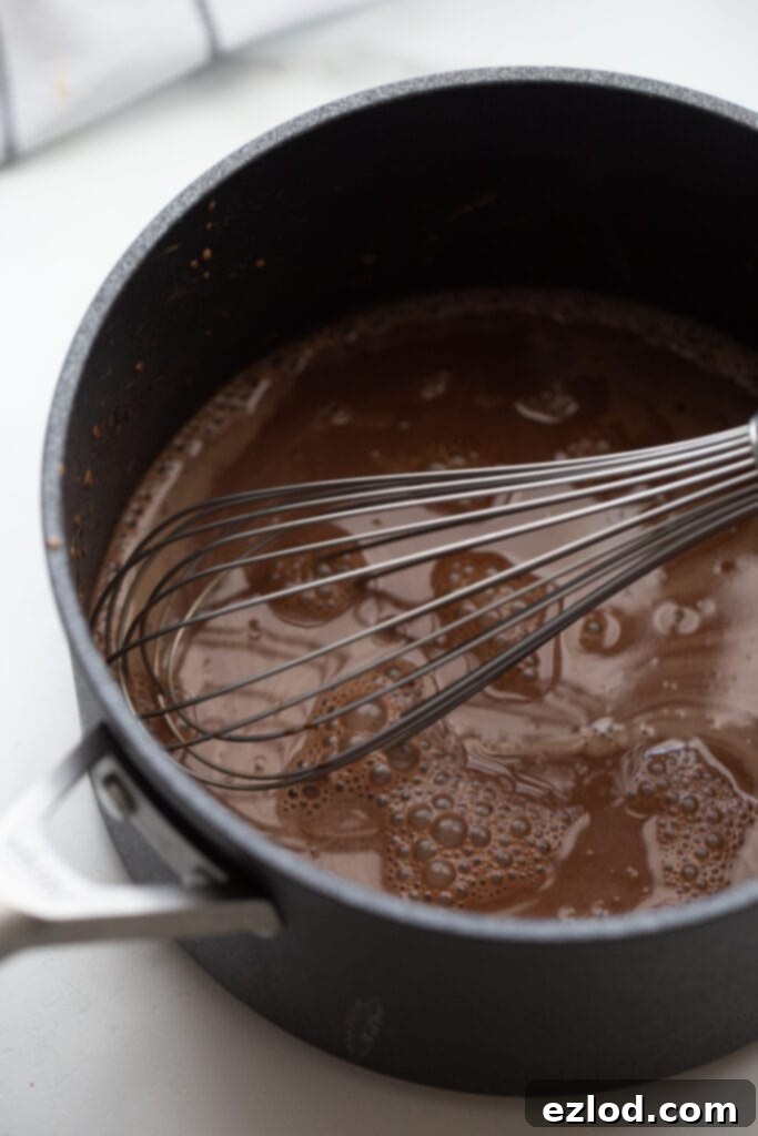 A hand whisking the homemade chocolate pudding mixture in a saucepan over medium heat, demonstrating the constant stirring needed for a smooth consistency.