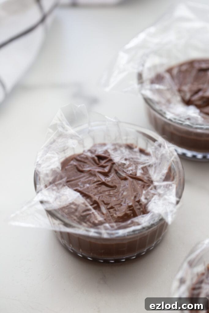 Small ramekins filled with smooth vegan chocolate pudding, each covered directly with plastic wrap to prevent a skin from forming while chilling.