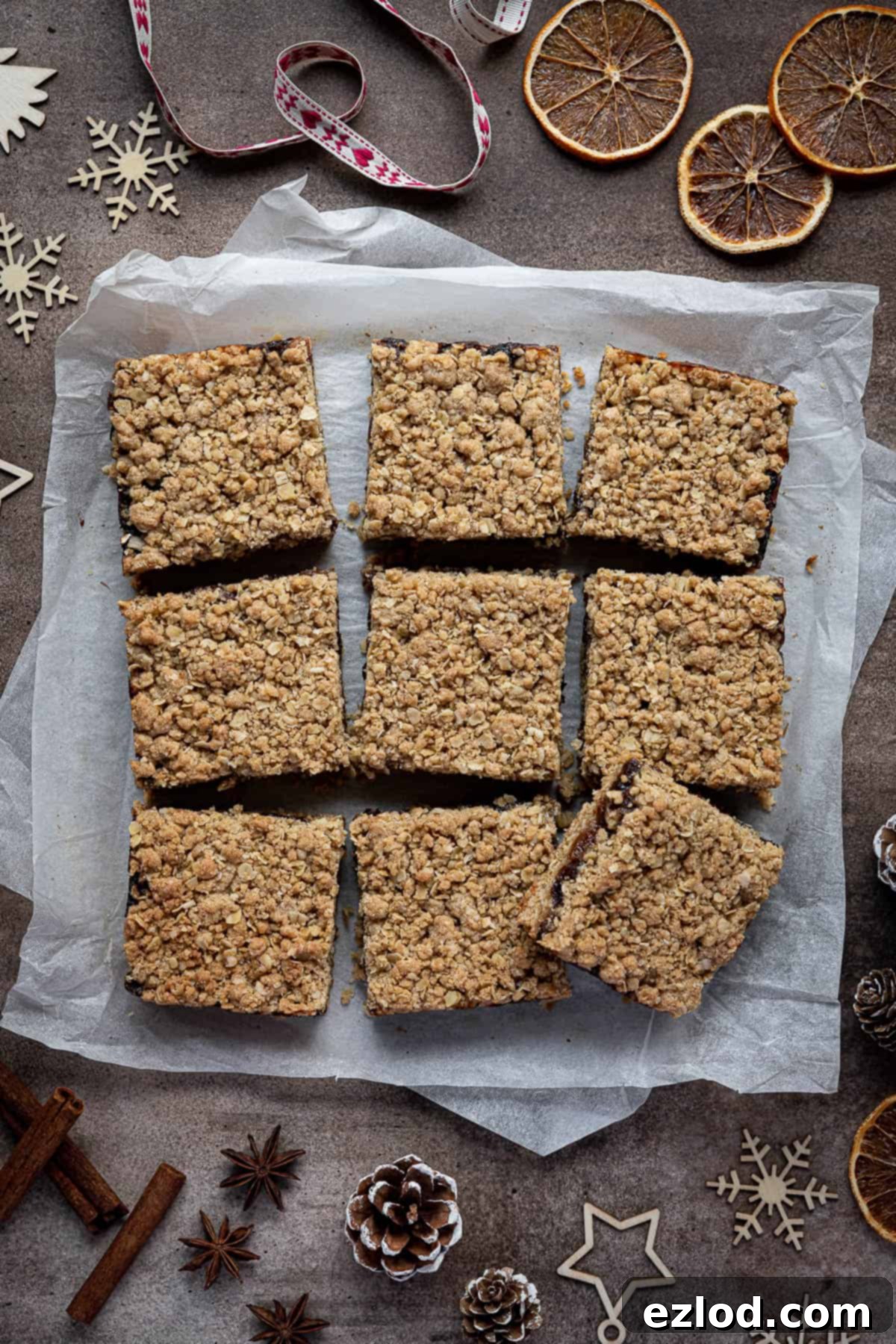 Vegan Mincemeat Crumb Bars 8 A beautifully arranged scene of vegan mincemeat crumble bars on baking parchment, surrounded by festive elements like cinnamon sticks, dried orange slices, and small Christmas ornaments, highlighting their holiday appeal.
