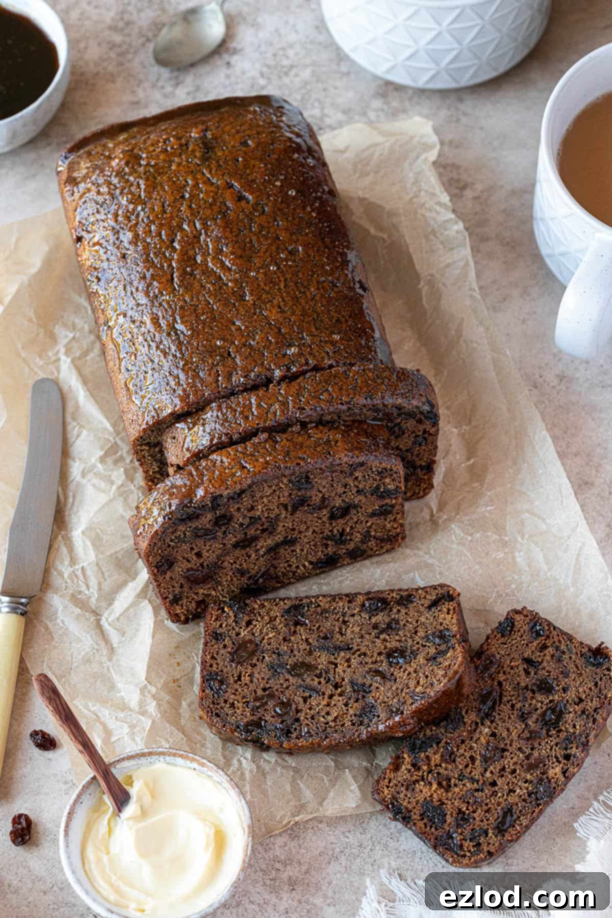 Sliced vegan malt loaf on a sheet of baking parchment with cups of tea and a bowl of butter.