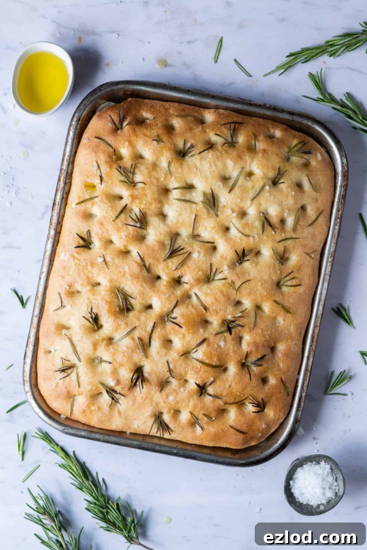 Golden brown sea salt and rosemary focaccia loaf in a baking tin on a marble background, surrounded by fresh rosemary sprigs, bowls of olive oil, and flaky sea salt.