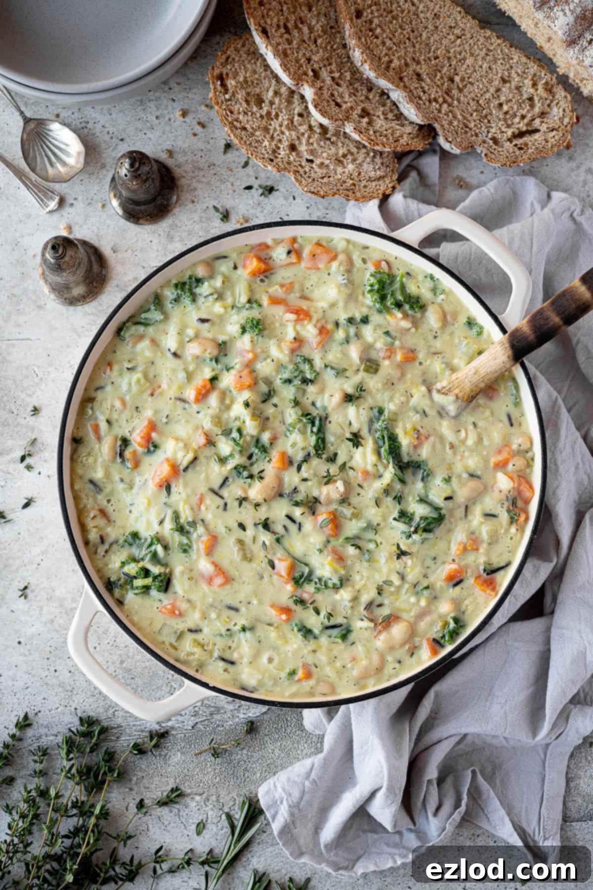 A beautifully composed flat lay image featuring a large pan of creamy vegetable rice soup, surrounded by fresh herbs, rustic bread slices, soup bowls, and salt and pepper shakers, highlighting a cozy meal setting.