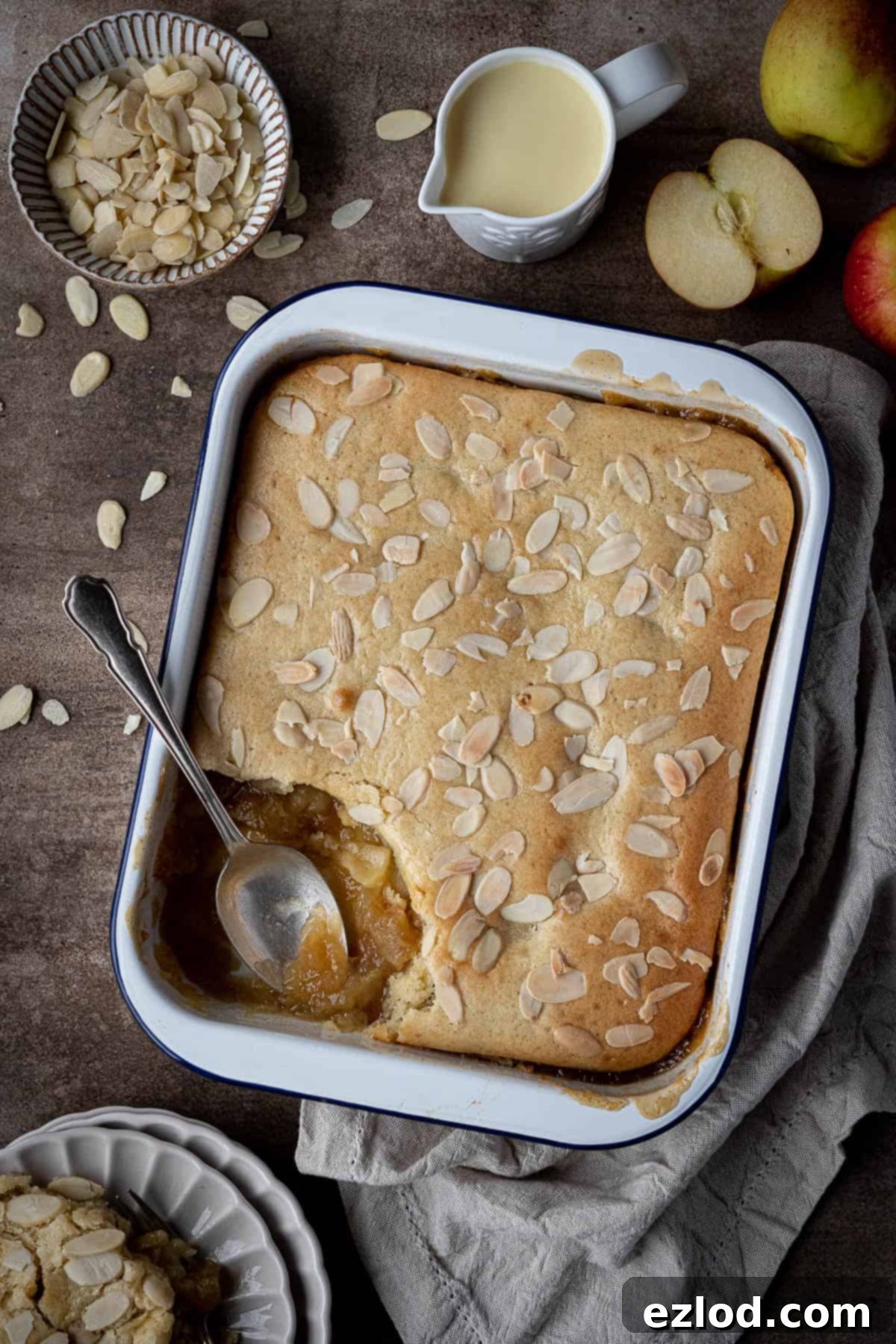 Delicious Vegan Eve's pudding in a white baking dish on a rustic brown surface, accompanied by a jug of vegan custard, a bowl of flaked almonds, and fresh apple slices. A perfect heartwarming dessert.