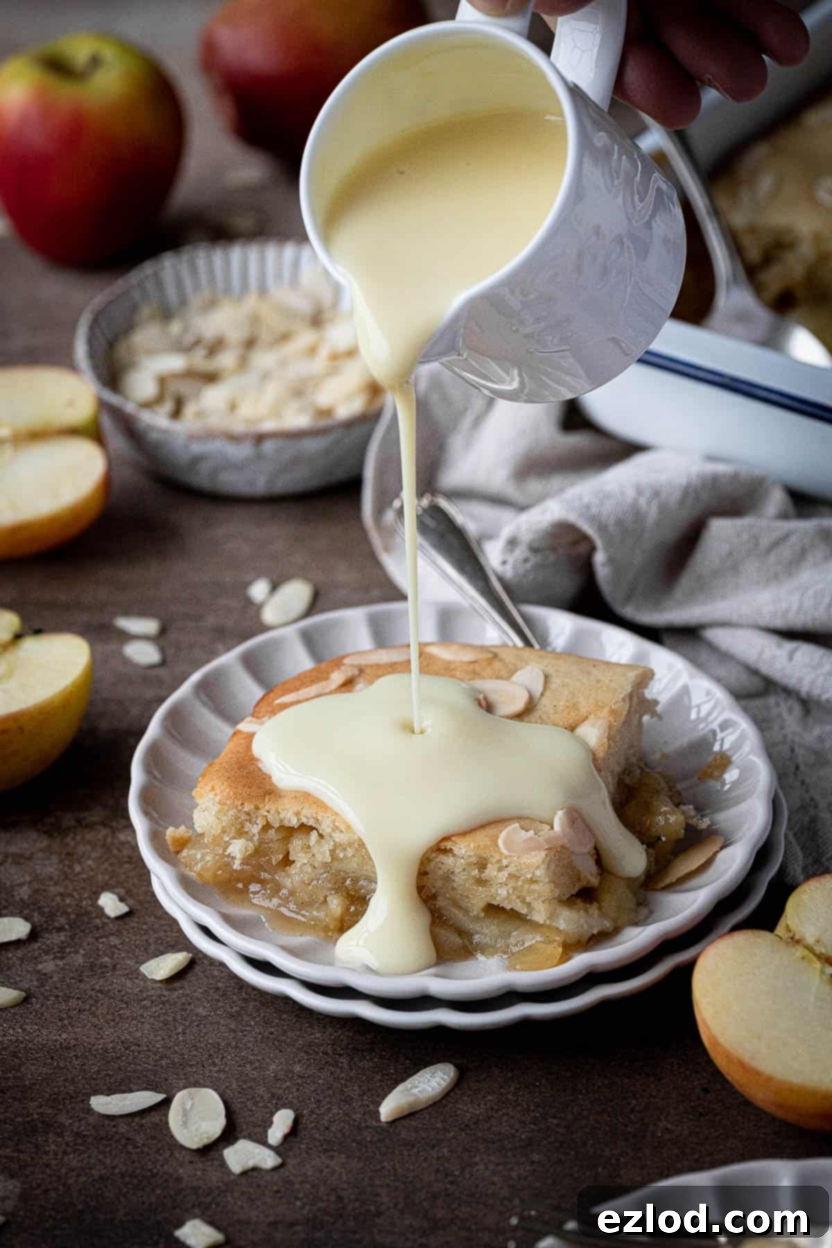 A stream of creamy vegan custard being poured over a generous slice of freshly baked Eve's pudding on a white plate, highlighting its comforting appeal.