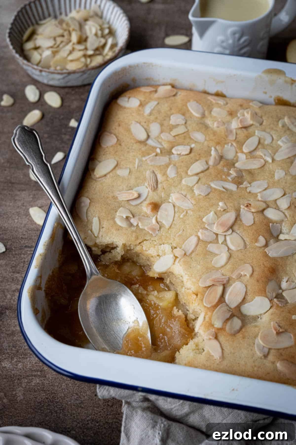 Close-up view of a freshly baked vegan Eve's pudding in a white baking dish with a spoonful carefully removed, showcasing the tender sponge and soft apple layer beneath.