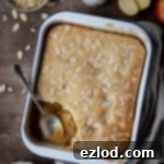 Vegan Eve's pudding in a white baking dish on a brown surface with a jug of custard, bowl of flaked almonds and sliced apples.