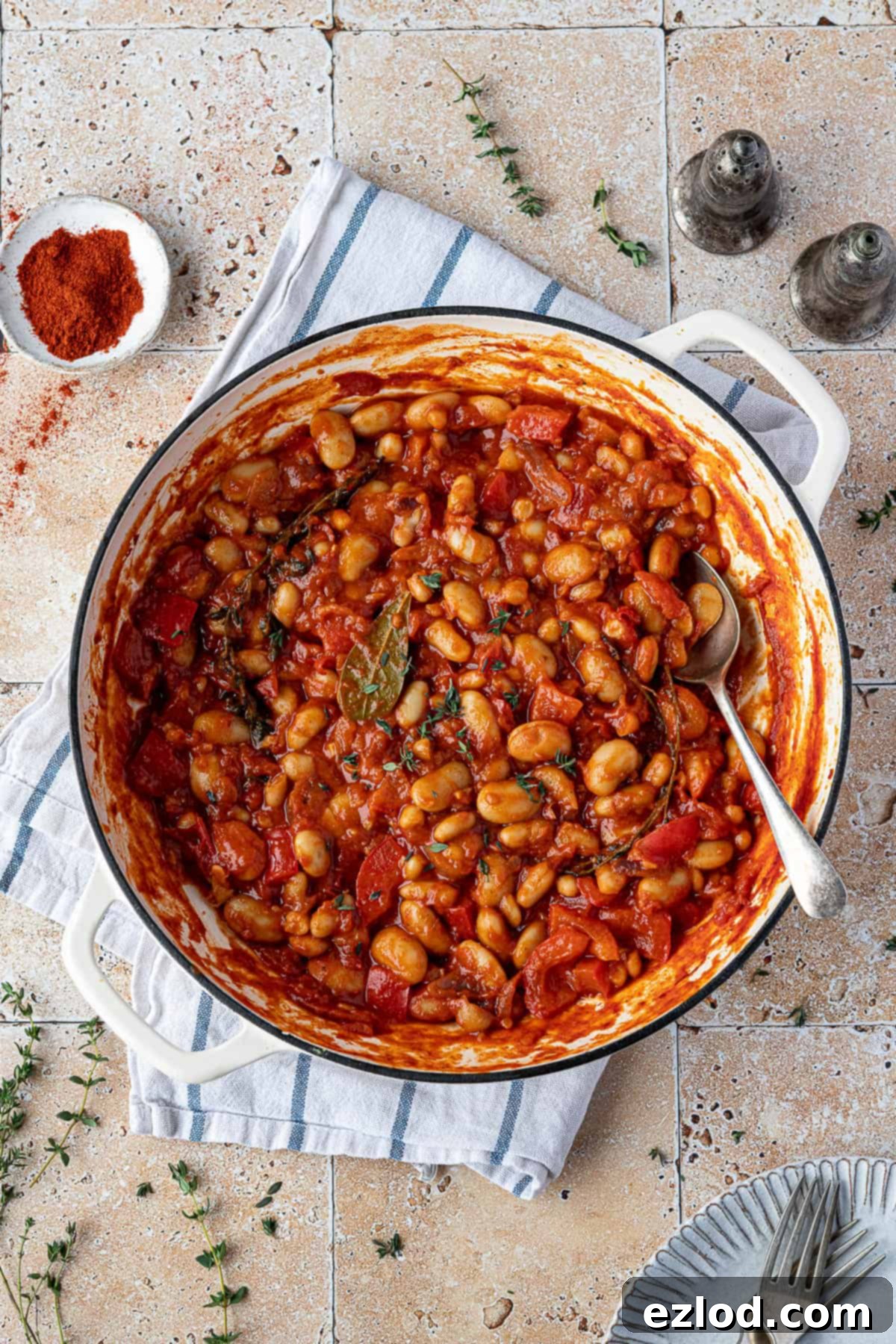 Hearty Smoky Tomato Beans 2 A rustic white pan brimming with rich, smoky tomato beans, garnished with fresh thyme. Surrounding it are a small bowl of vibrant paprika, and salt and pepper shakers.