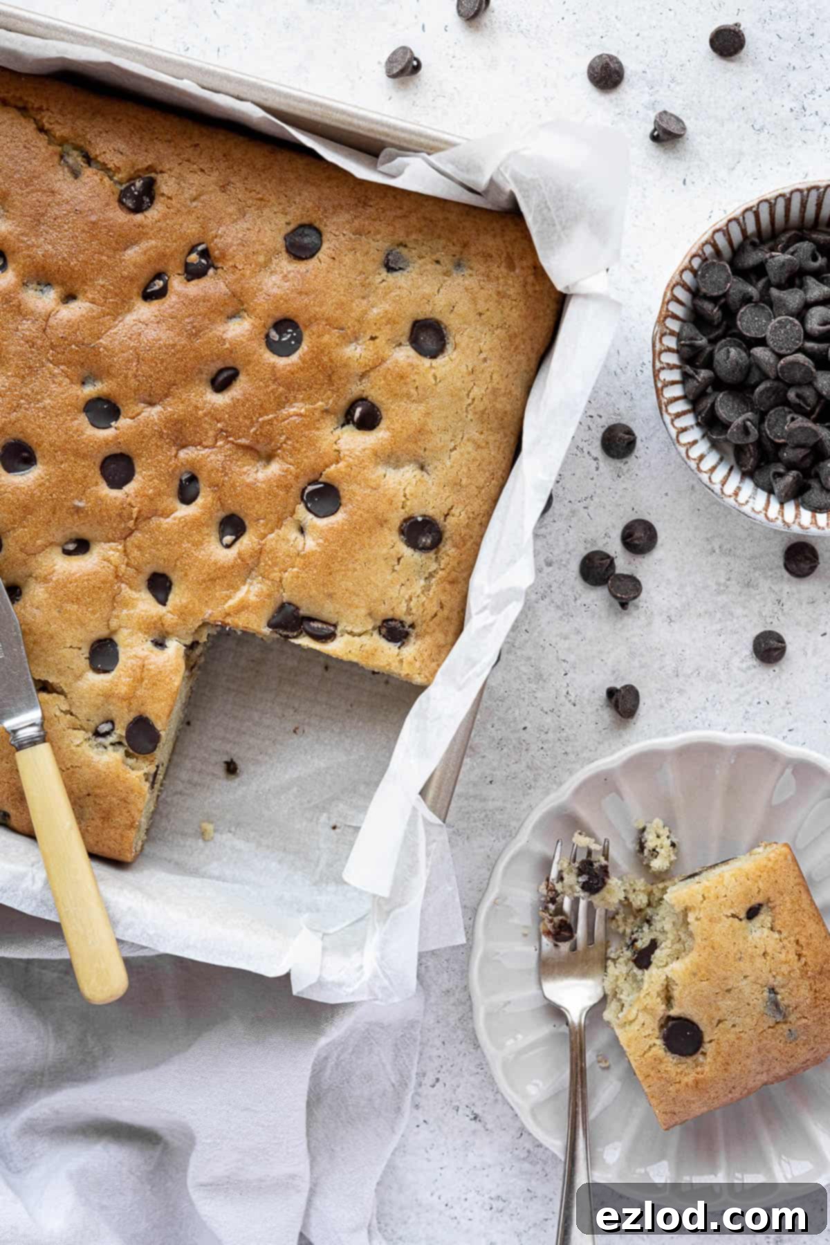 A square tin holds a freshly baked vegan chocolate chip vanilla cake, with one slice elegantly removed, revealing its moist interior.