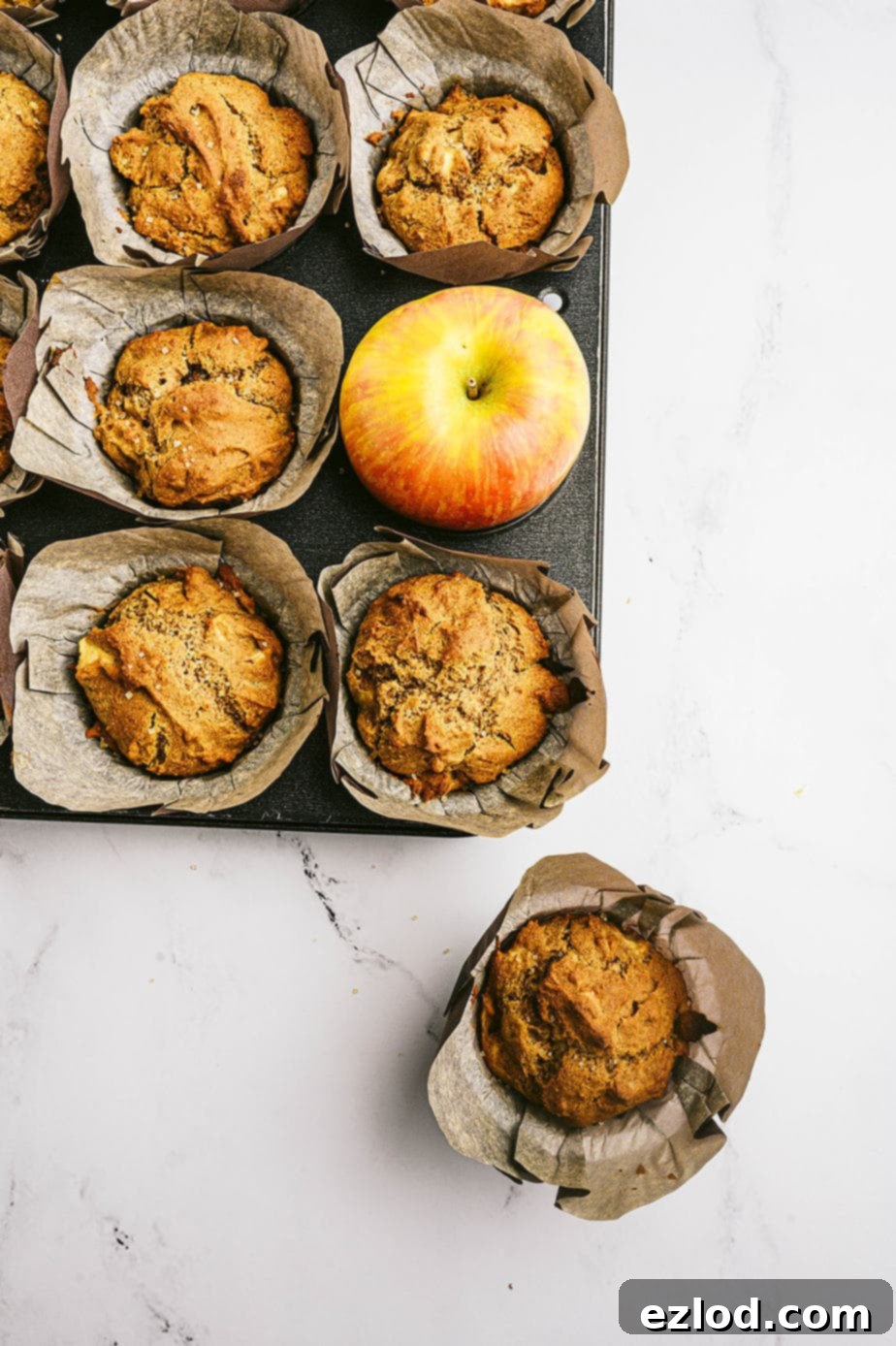 Overhead image: parchment lined muffins in a muffin tin. One muffin is out of its place, off to the side and a whole apple takes its place in the tin.