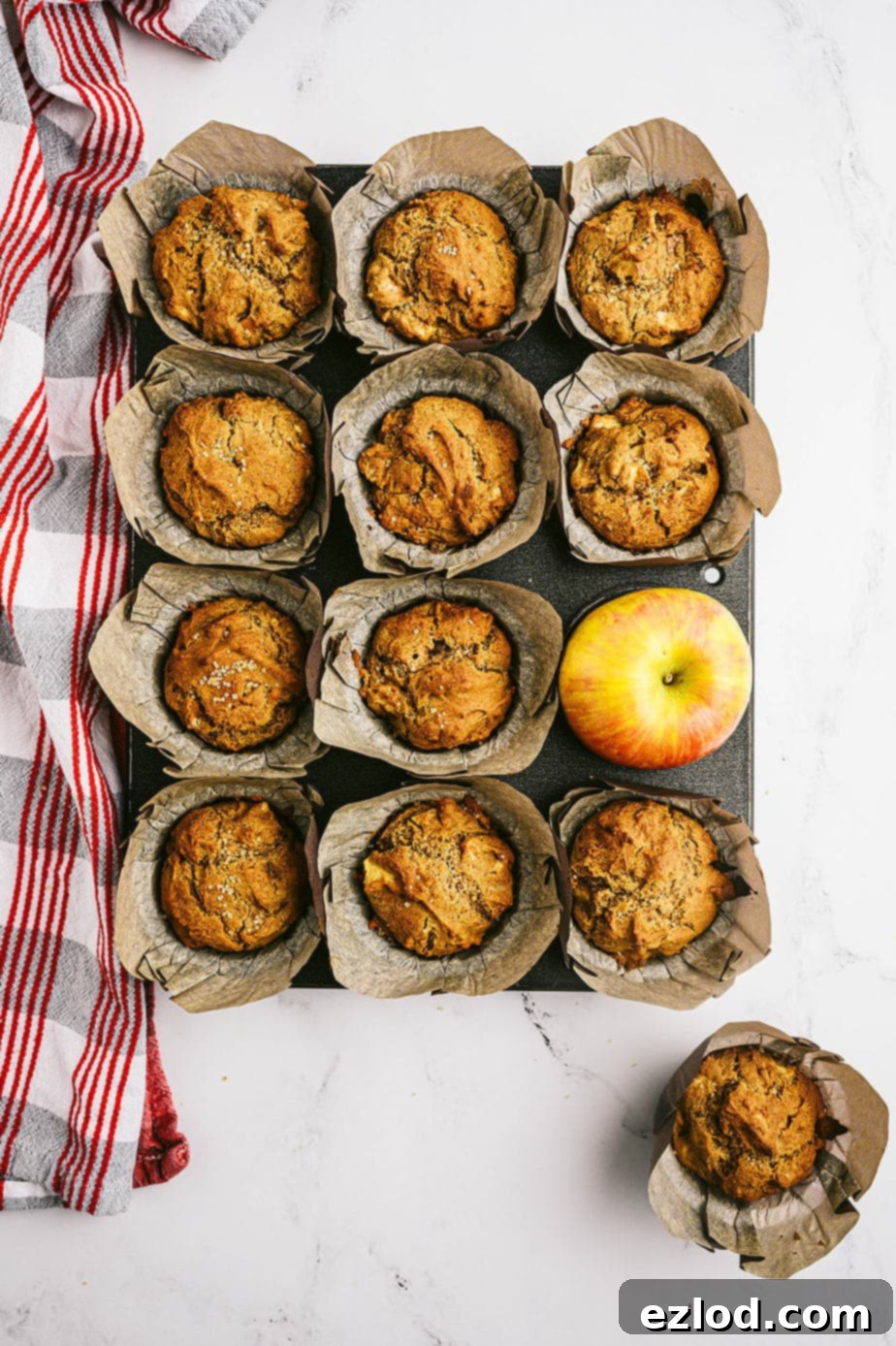 Overhead photo: muffin tin full of cooked muffins with a whole apple taking one's place.