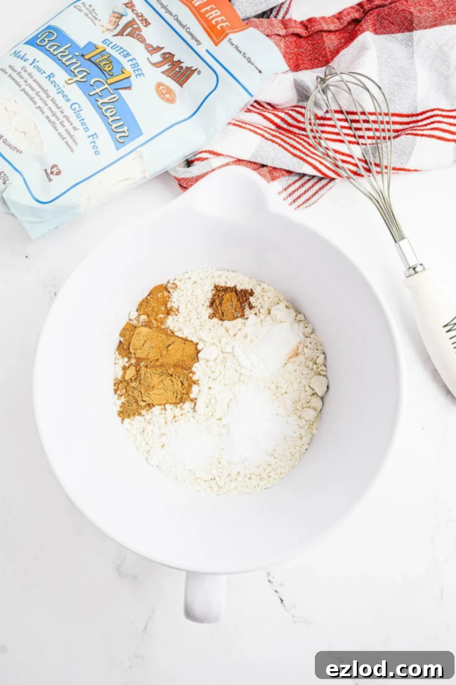Overhead photo of dry ingredients in a white bowl. A bag of Bob's Red Mill 1 to 1 Baking Flour and a whisk are laying off to the side.