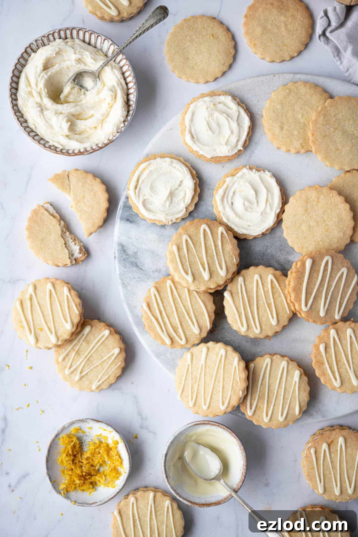 Vegan lemon sandwich cookies on a marble board with bowls of buttercream, lemon zest and white chocolate.