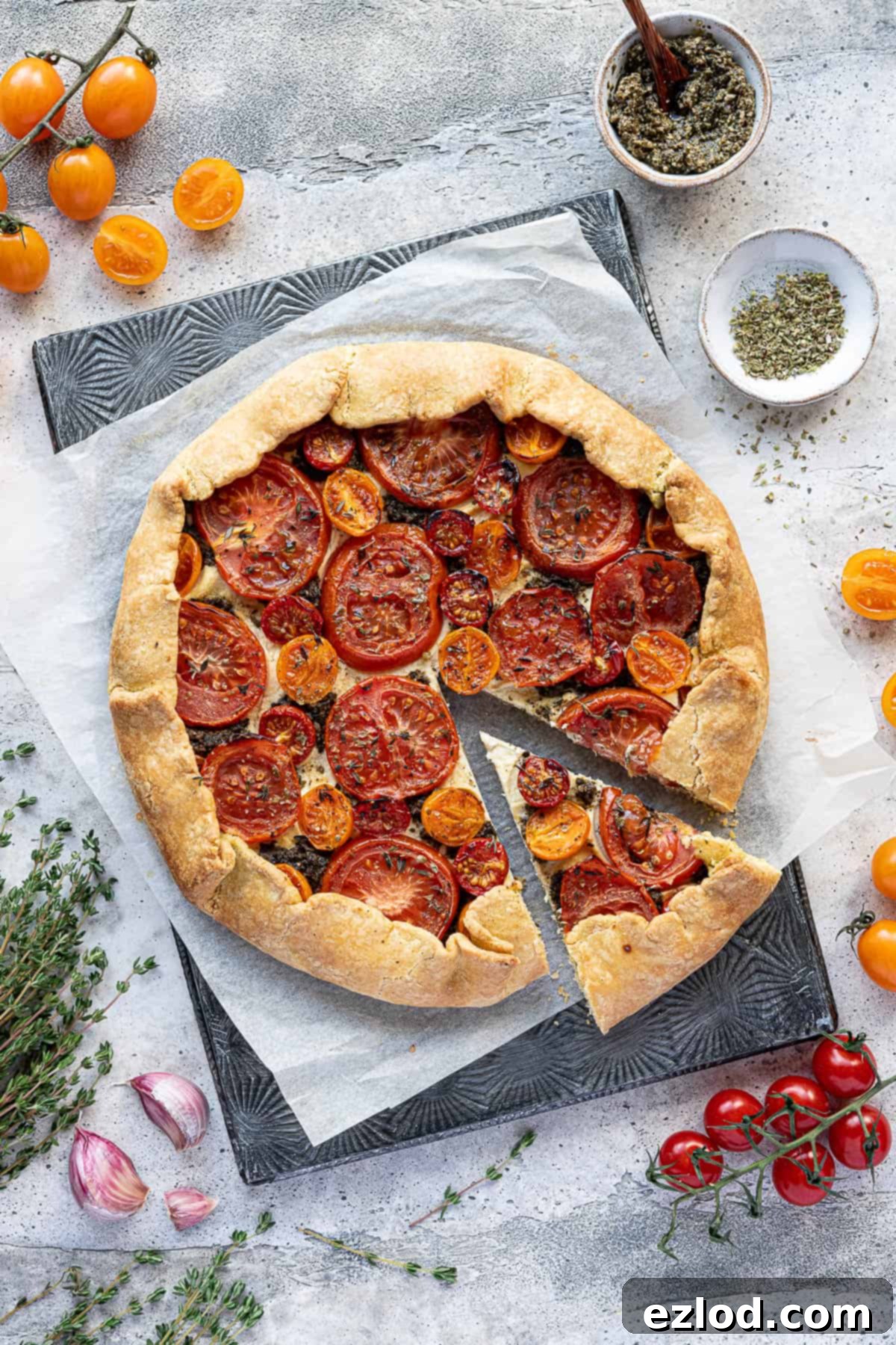A perfectly baked vegan tomato ricotta galette with a slice cut out, resting on baking parchment. It is surrounded by fresh tomatoes, herbs, and a small bowl of green pesto, highlighting its vibrant ingredients.