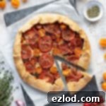 Vegan tomato ricotta galette with a slice cut out on a sheet of baking parchment surrounded by tomatoes, herbs and a bowl of pesto.