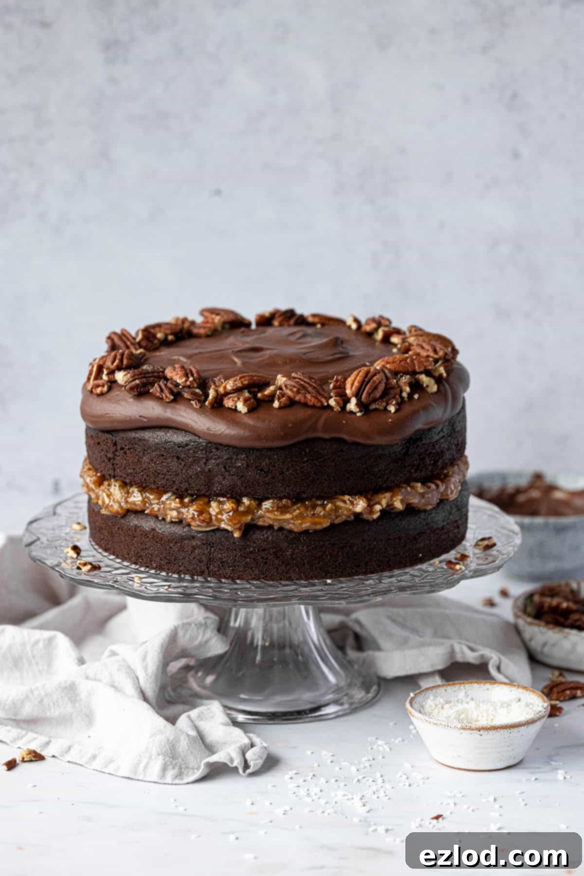 Vegan German chocolate cake on a glass cake stand with bowls of pecans, coconut and ganache.