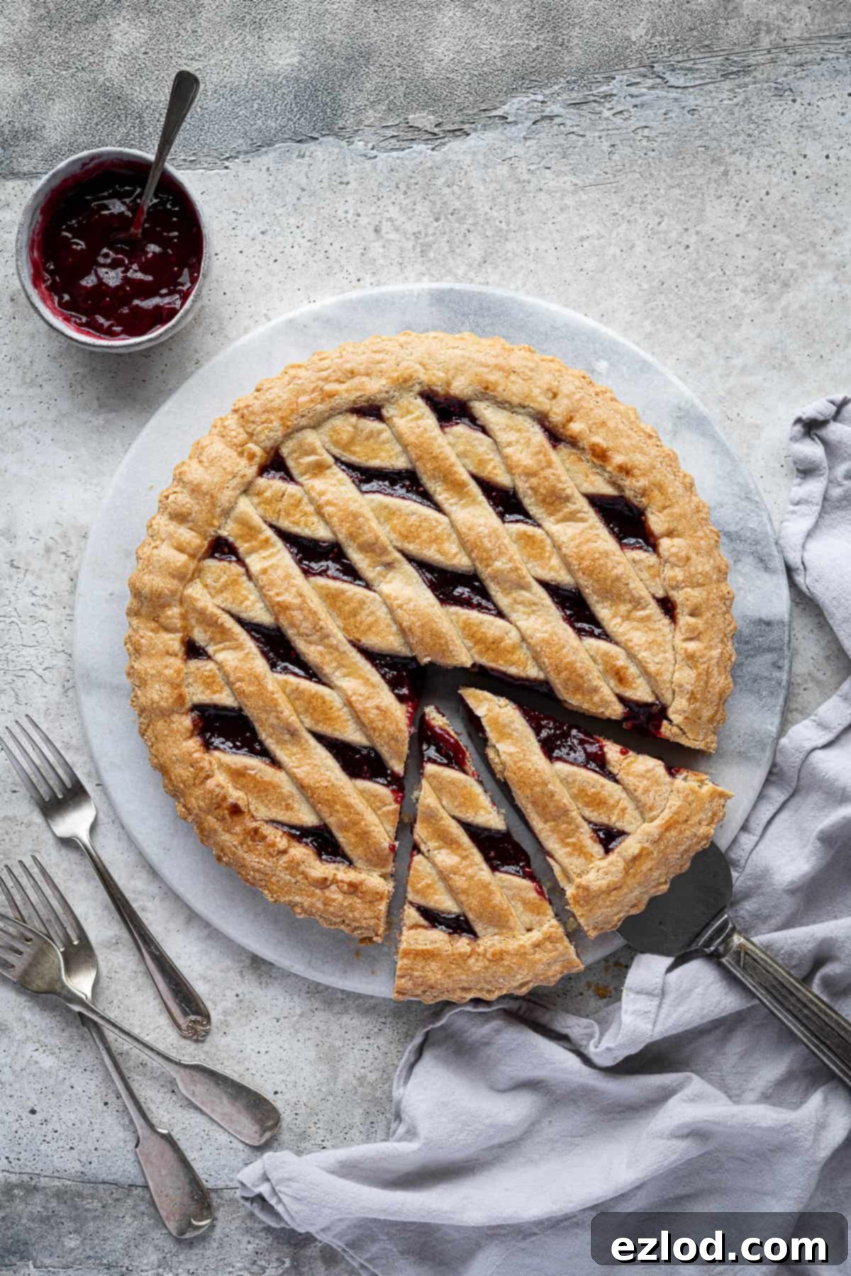 Beautifully baked vegan jam crostata on a white marble board, garnished with fresh berries and a small bowl of raspberry jam. Forks are placed beside it, ready to serve.