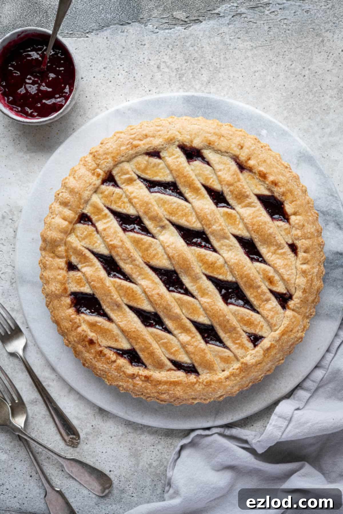 A freshly baked vegan jam crostata cooling on a white marble board, beautifully golden-brown with a glistening jam filling, sitting beside a small bowl of raspberry jam. The scene is inviting and delicious.
