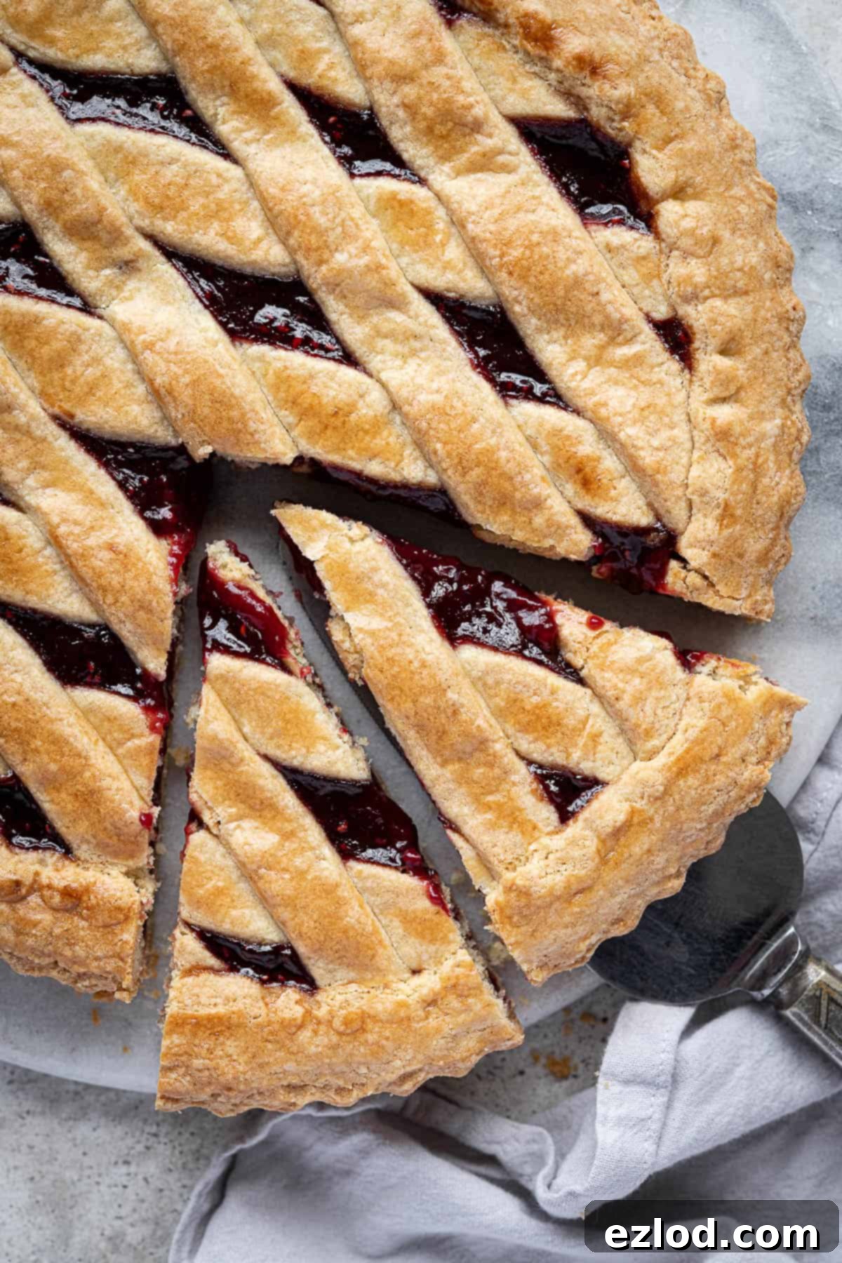 Close-up view of a freshly baked vegan crostata with two slices already cut, revealing the vibrant raspberry jam filling and the perfectly golden, flaky pastry crust. The texture is inviting and delicious.