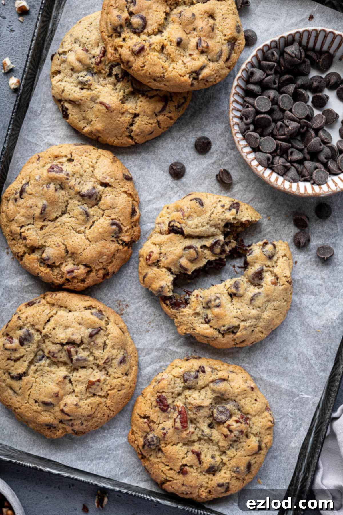 A baking tray filled with baked chunky chocolate pecan cookies, with one cookie broken in half to show its soft interior.
