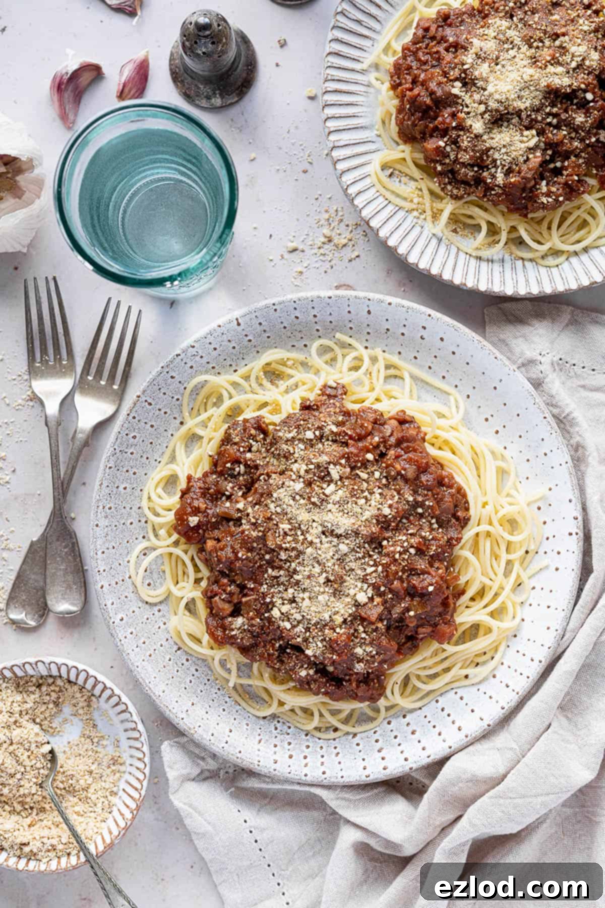 Two plates of vegan spaghetti bolognese with a glass of water and a bowl of vegan parmesan.