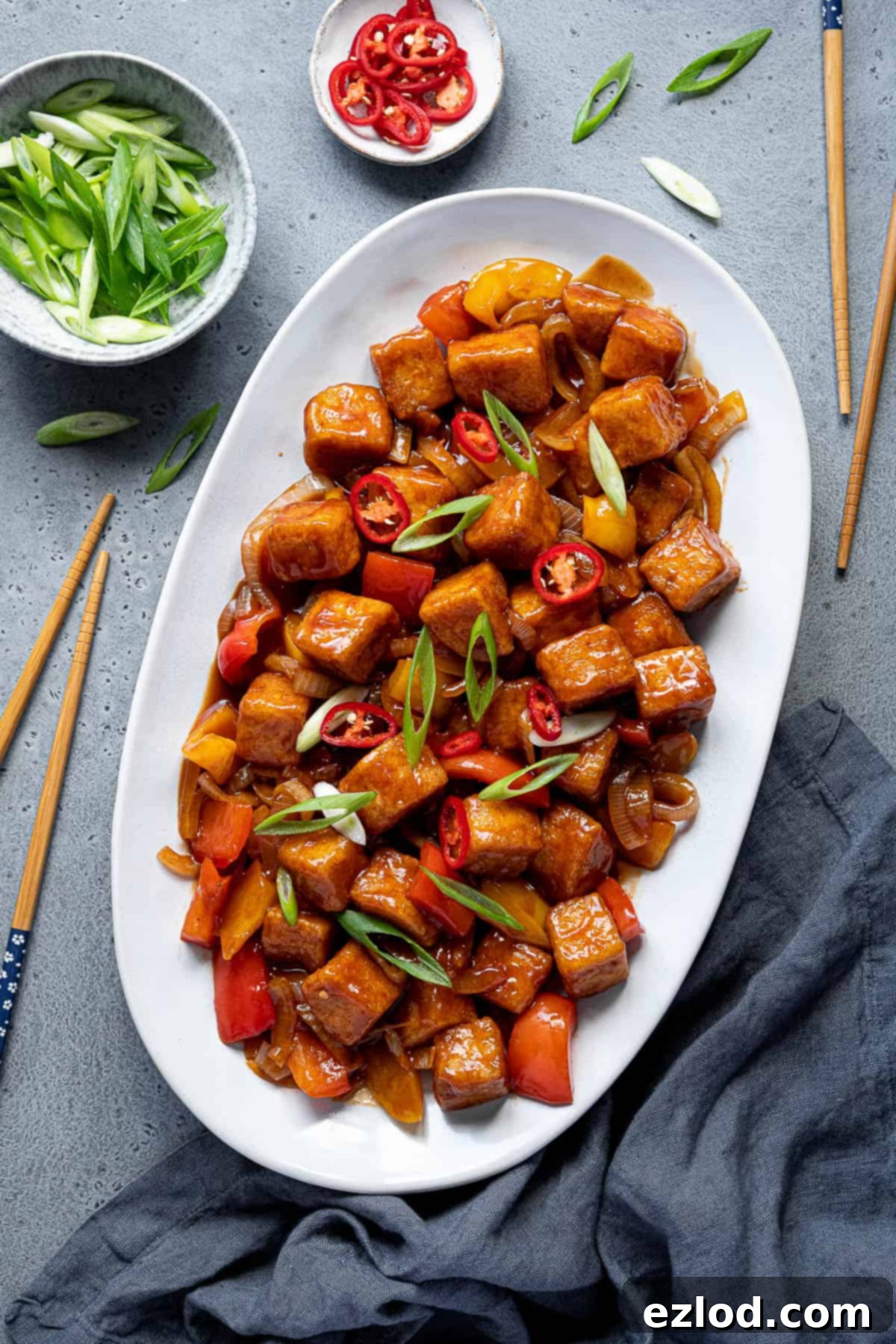 Sweet and sour tofu served on an oval white plate with chopsticks, garnished with fresh spring onion and chili flakes, alongside small bowls of extra toppings.