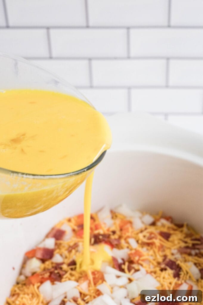 Pouring whisked egg mixture over layered vegetables, bacon, and cheese in a slow cooker