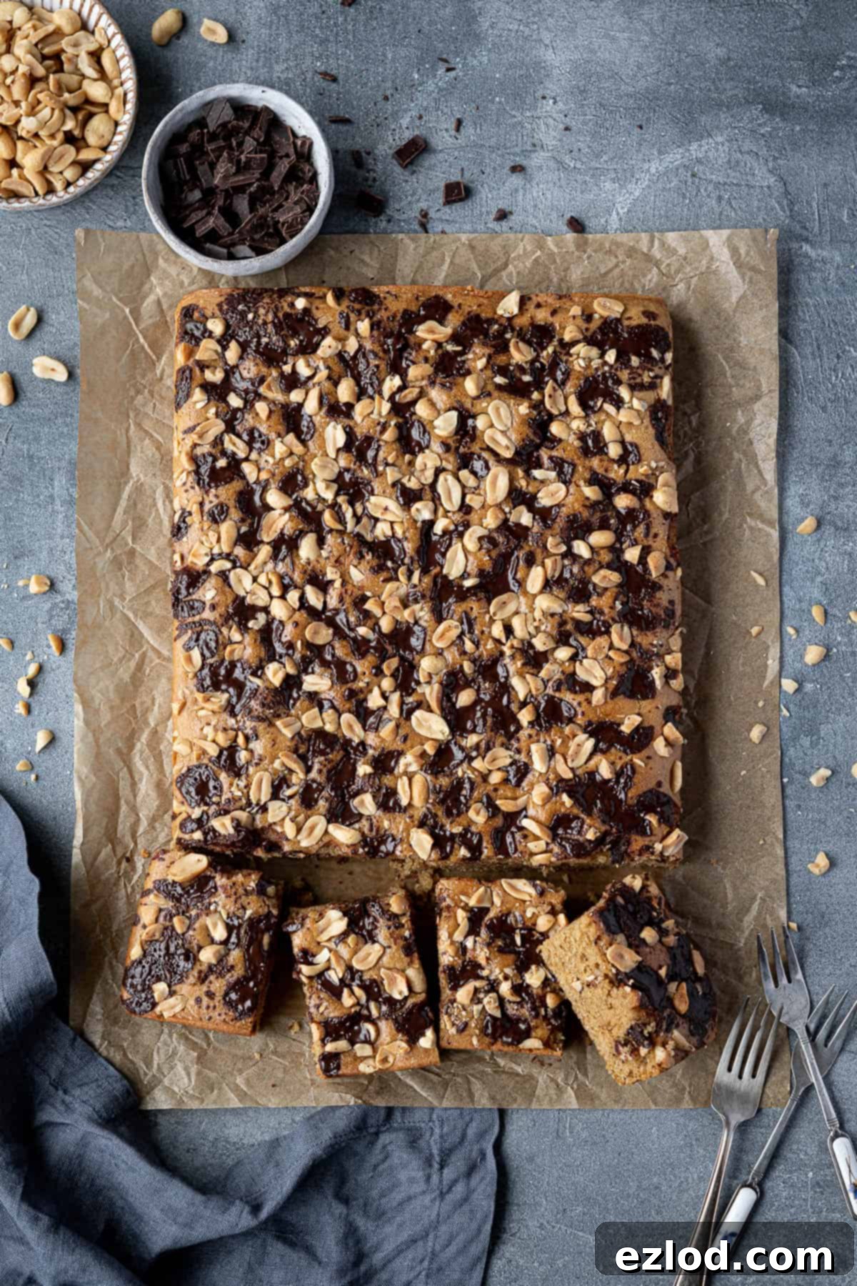 A freshly baked vegan peanut butter cake on baking parchment, with bowls of chocolate chips and roasted peanuts ready for topping.