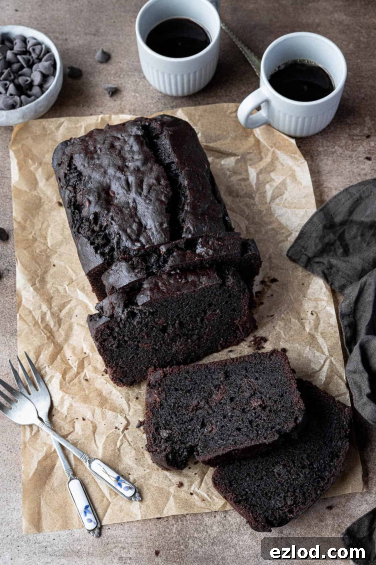 Sliced vegan chocolate courgette cake on a sheet of brown baking paper with cups of coffee, bowl of chocolate chips and cake forks.