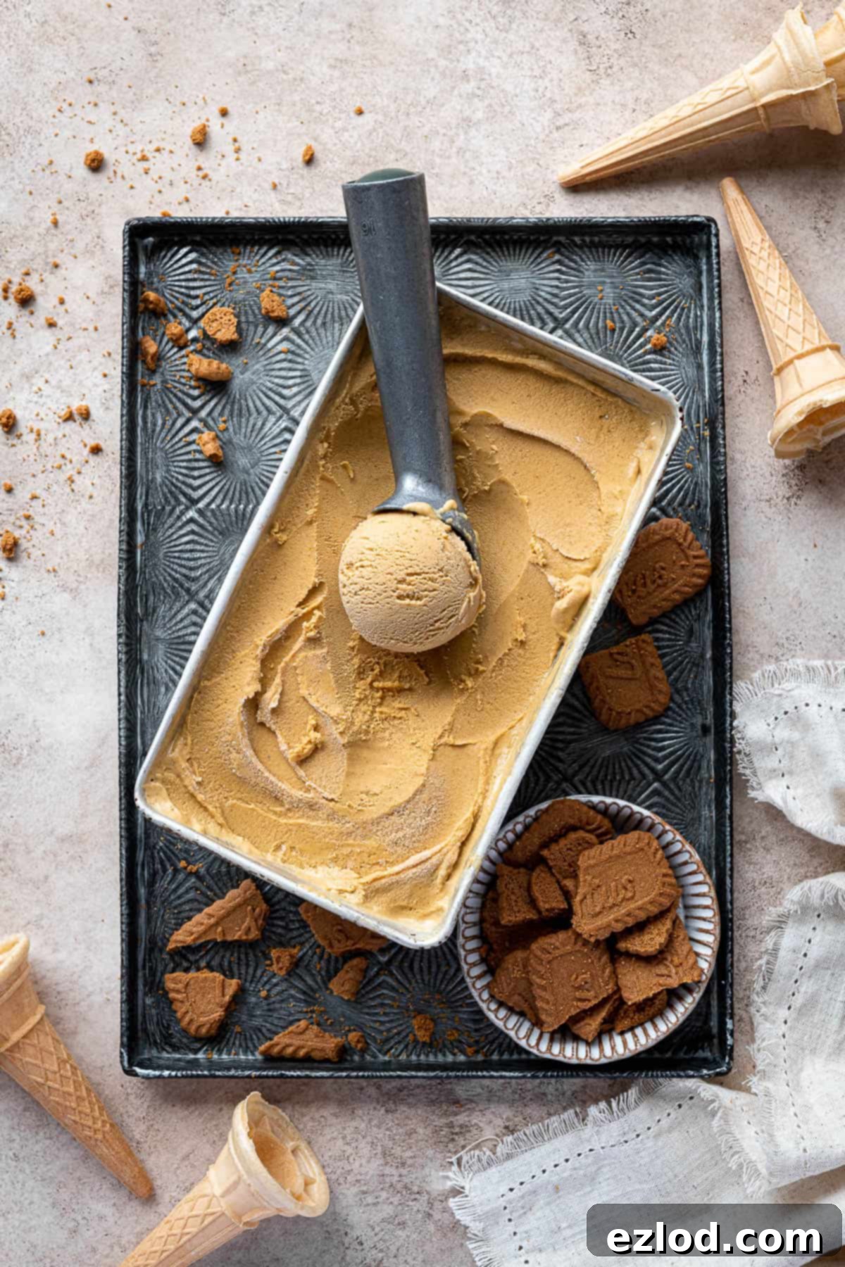 Vegan pumpkin ice cream in a loaf tin on a metal tray surrounded by ice cream cones and crushed cookies.